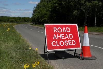 Red road closed sign across road with wildflower verges with traffic cone next to the sign